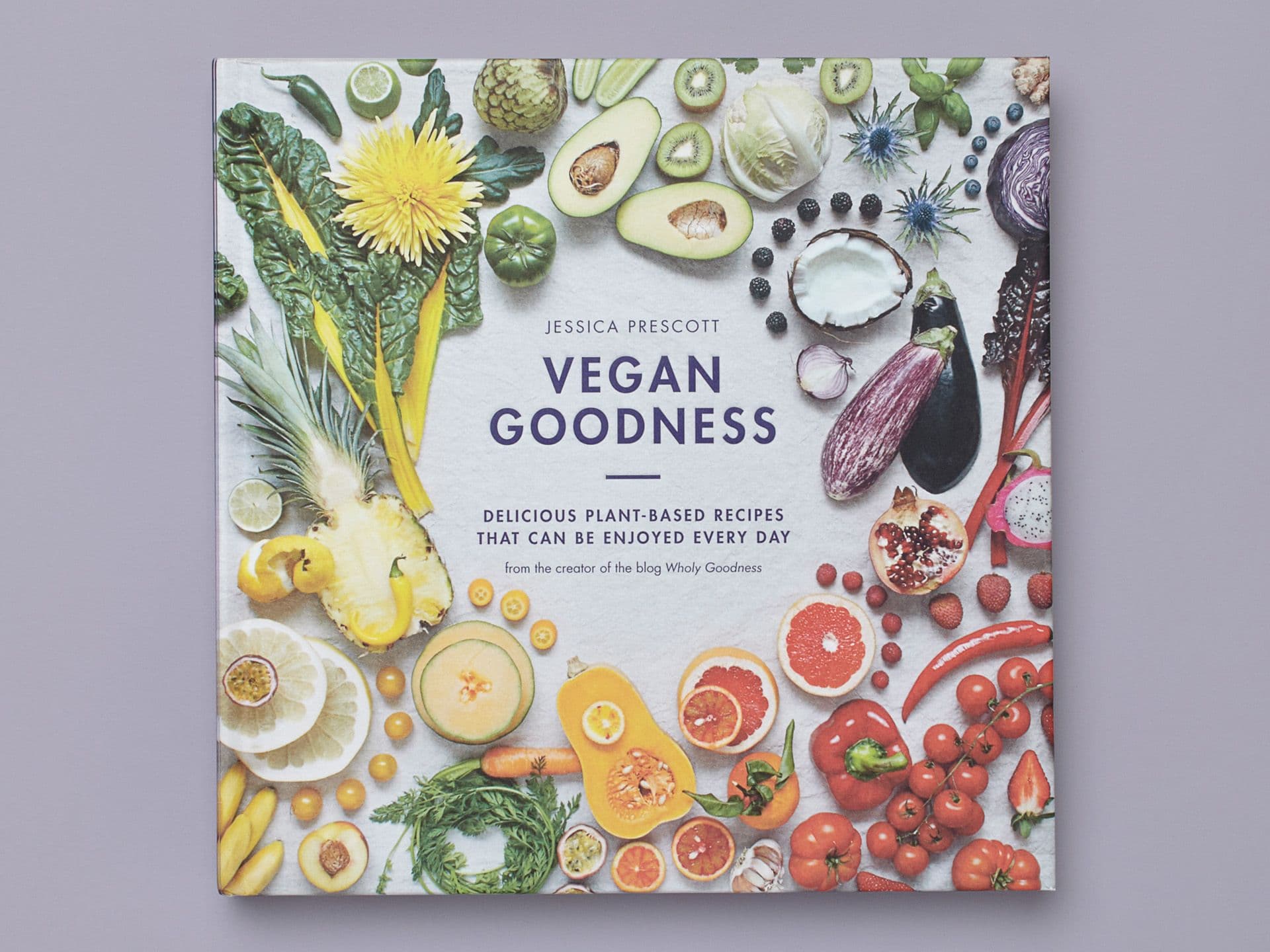 Book titled "Vegan Goodness" on a pale grey background, featuring a flat-lay of colourful fruits, vegetables, and edible flowers arranged around the cover. Subtitle reads "Delicious plant-based recipes that can be enjoyed every day," with the author's name, Jessica Prescott, centred at the top.