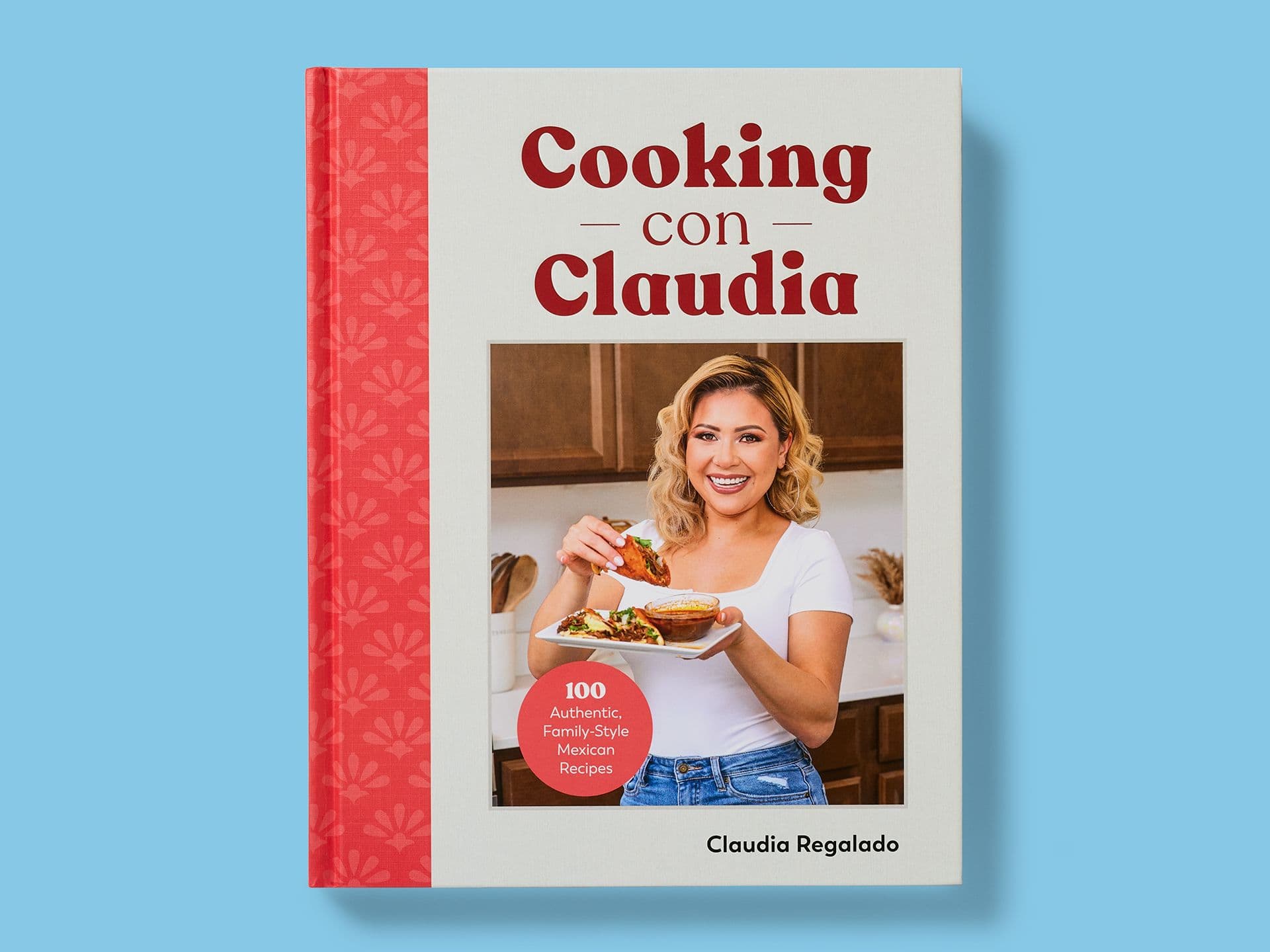 Book cover titled "Cooking con Claudia" by Claudia Regalado, featuring a portrait of the author holding a plate of Mexican food, a red spine with a floral pattern, and a circular badge reading "100 Authentic, Family-Style Mexican Recipes," set against a light blue background.