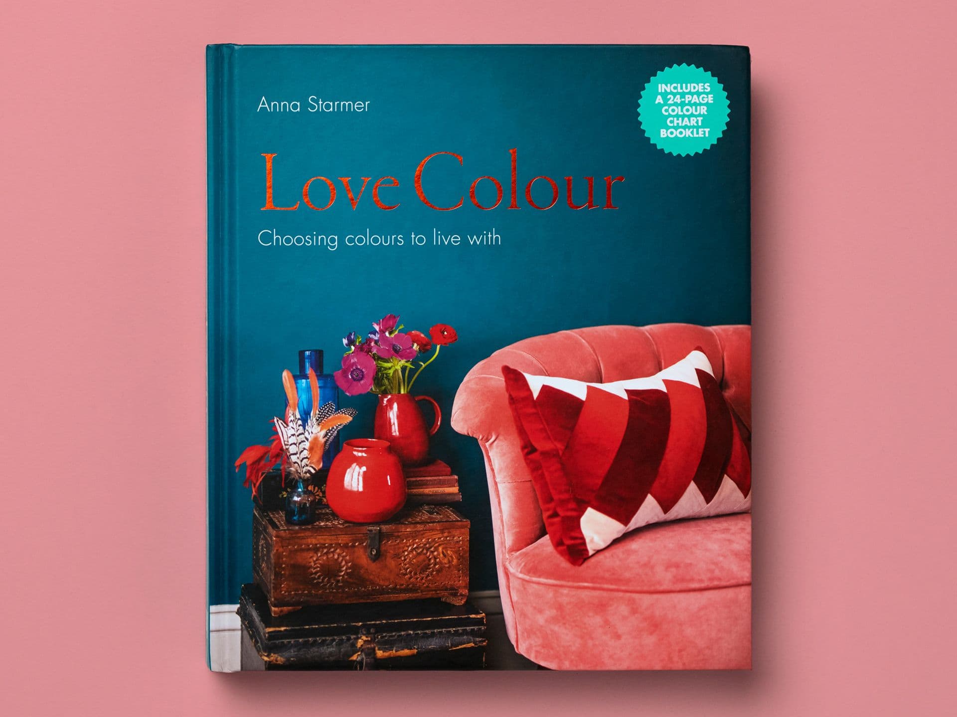 Book cover titled "Love Colour" by Anna Starmer on a pink background, featuring a teal wall, a pink velvet armchair with a geometric cushion, and a side table with red vases and flowers. Subtitle reads "Choosing colours to live with," with a circular badge stating "Includes a 24-page colour chart booklet."