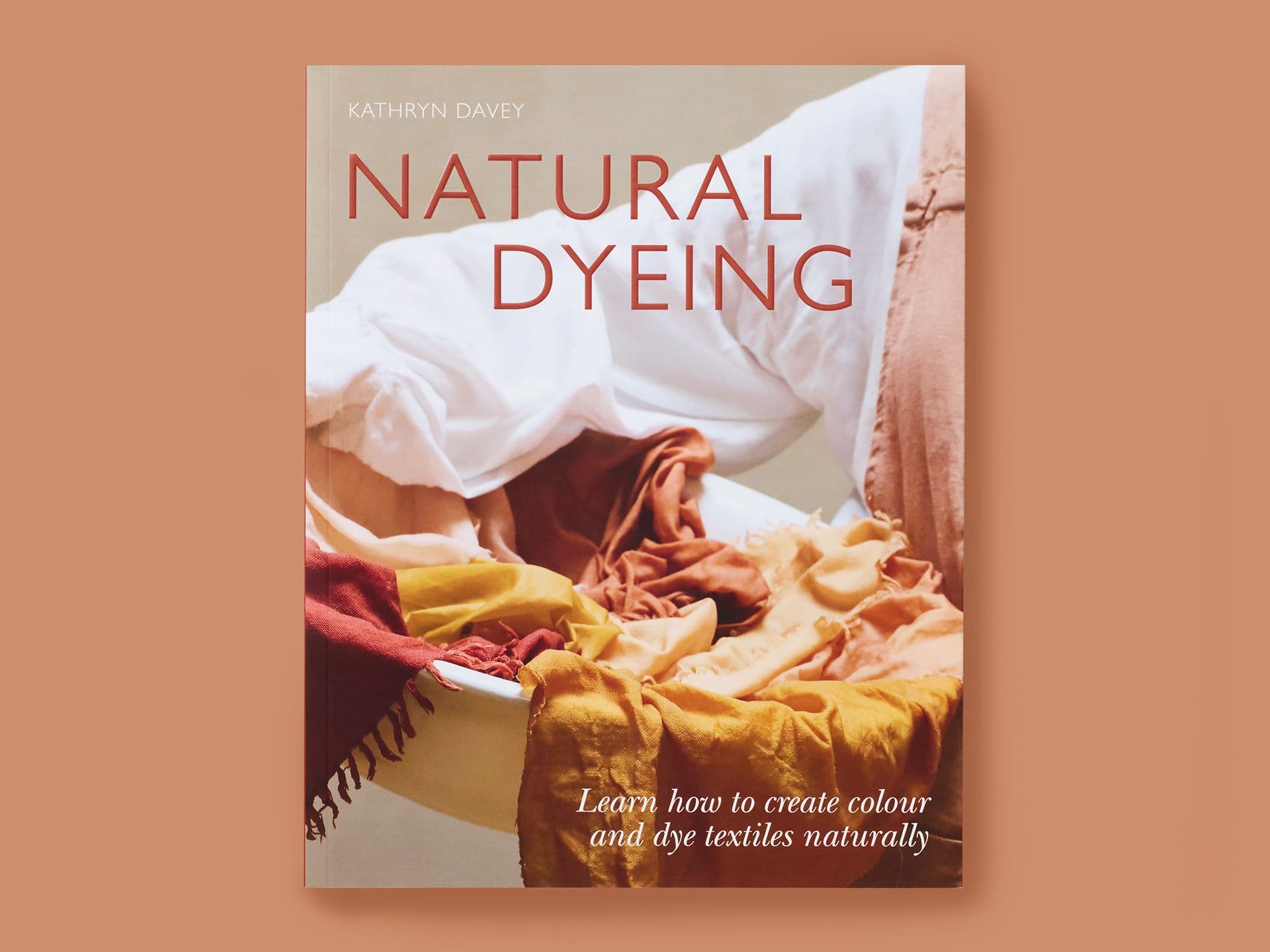 Book titled "Natural Dyeing" on a warm terracotta background, featuring softly draped textiles in earthy tones of rust, ochre, and cream arranged in a basin. Subtitle reads "Learn how to create colour and dye textiles naturally," with the author's name, Kathryn Davey, at the top.