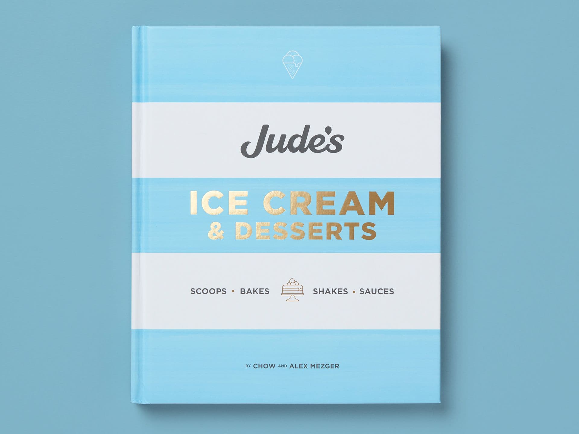 Book titled "Jude’s Ice Cream & Desserts" on a pale blue background, with soft horizontal stripes and gold-foil lettering. Subheading lists "Scoops, Bakes, Shakes, Sauces," with a small dessert icon and author credit to Chow and Alex Mezger at the bottom.