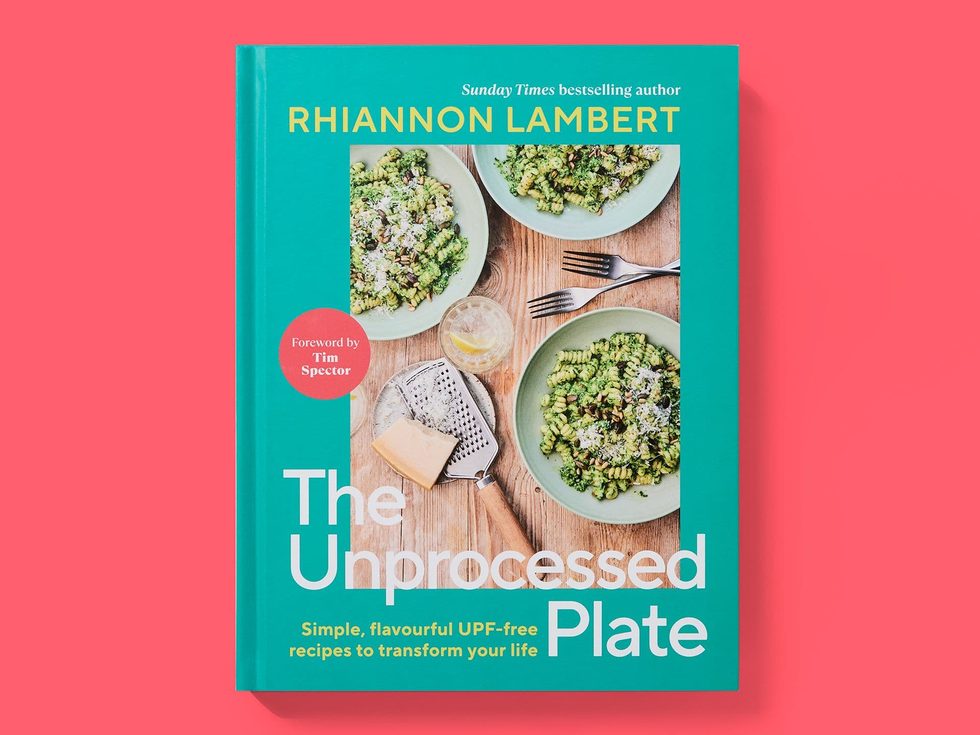 Teal book cover titled "The Unprocessed Plate" by Rhiannon Lambert, featuring a food photograph of pasta dishes, forks, cheese, and a grater on a wooden table, with the subtitle "Simple, flavourful UPF-free recipes to transform your life" and a red badge noting a foreword by Tim Spector, set against a pink background.